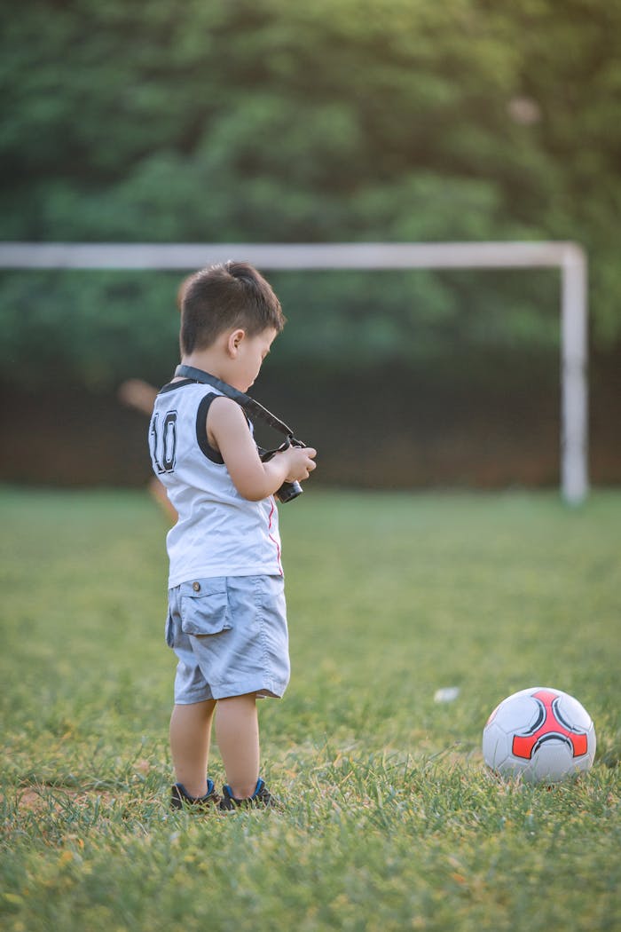 A young boy stands on a soccer field with a ball, enjoying a sunny day outdoors.