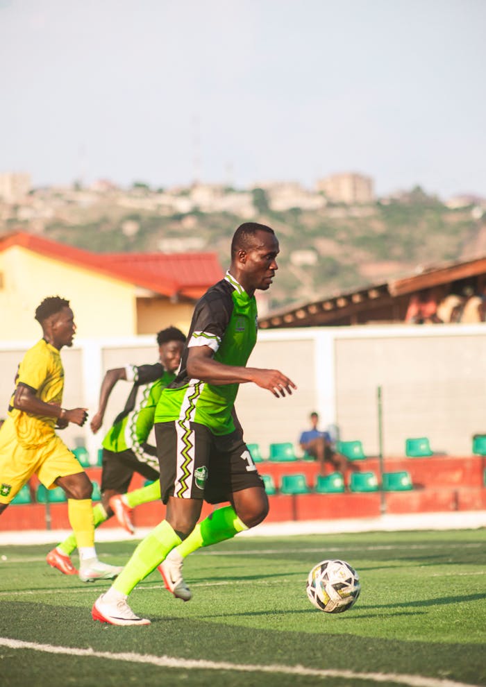 Home Youth football match with players in vibrant uniforms running on field under sunny sky.